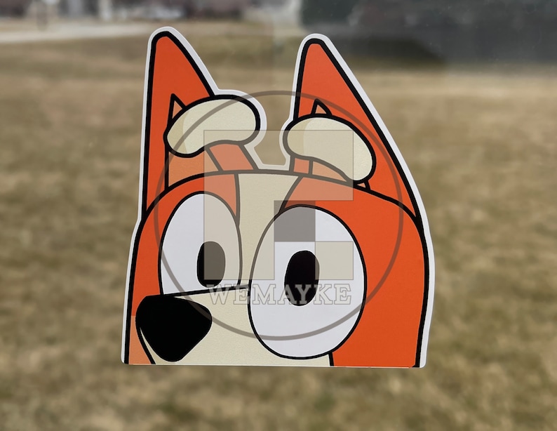 May include: A die-cut vinyl sticker featuring a cartoon dog character with orange fur, cream-colored accents, and large white eyes with black pupils. The character has pointed ears and a black nose. The text WEMAYKE is visible in the center.