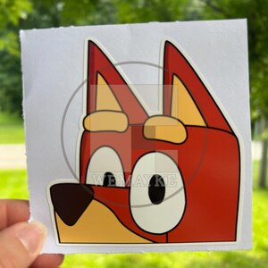 May include: A square sticker featuring a cartoon dog character with red fur, large white eyes with black pupils, a dark brown nose, and yellow inner ears. The character has pointed ears and a light yellow muzzle. The text WEMAYKE is visible.