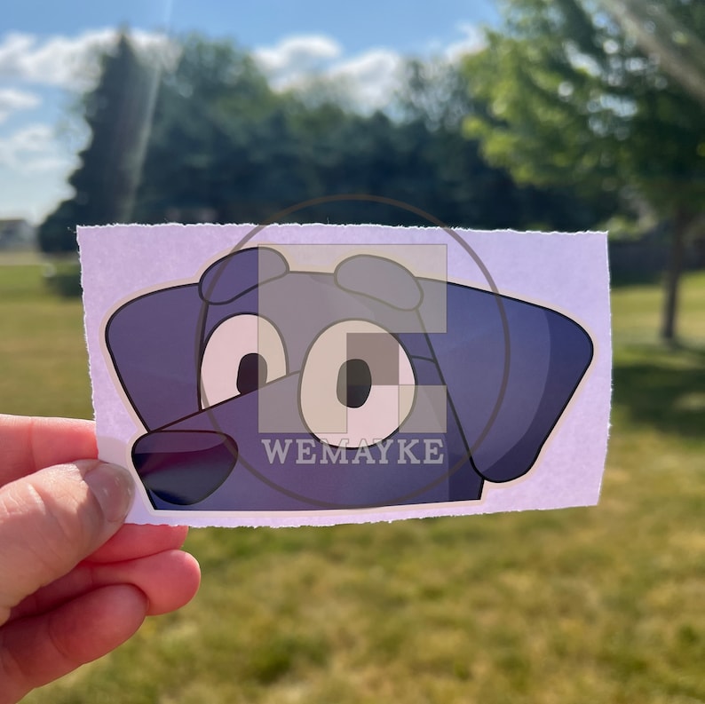 May include: A rectangular sticker featuring a stylized cartoon dog head with large white eyes, dark blue fur, and a black nose. The design includes a semi-transparent overlay with the text WEMAYKE centered over the dog's face on a white background.