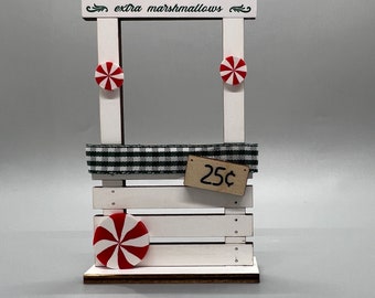 May include: A white wooden hot cocoa stand with a red and green sign that says "HOT COCOA extra marshmallows". There are two red and white peppermint candies on the stand, and a sign that says "25¢".
