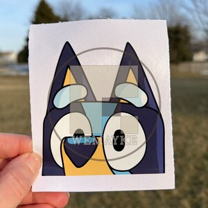 May include: A square sticker featuring a graphic illustration of a blue dog character with large white eyes, dark blue ears, and a light yellow snout. The design includes a semi-transparent overlay with the text WEMAYKE centered on the image.