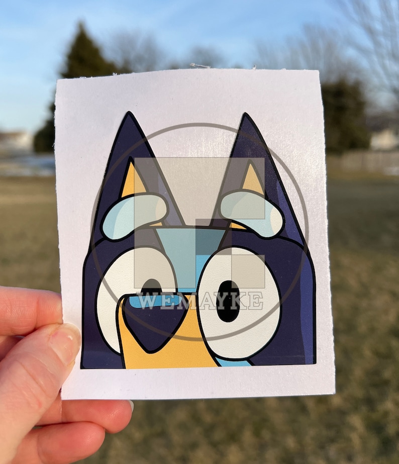 May include: A square sticker featuring a graphic illustration of a blue dog character with large white eyes, dark blue ears, and a light yellow snout. The design includes a semi-transparent overlay with the text WEMAYKE centered on the image.