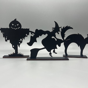 May include: Black Halloween-themed wooden cutouts. The set includes a scarecrow, a witch on a broom, and a cat with an arched back. Each cutout is mounted on a small, rectangular base. The figures are all black.