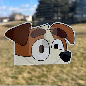 May include: A die-cut vinyl sticker featuring a cartoon dog head with brown ears, brown eyebrows, and large white eyes with dark pupils. The dog has a white muzzle and a black nose. The text WEMAYKE is visible in a faint watermark across the center.