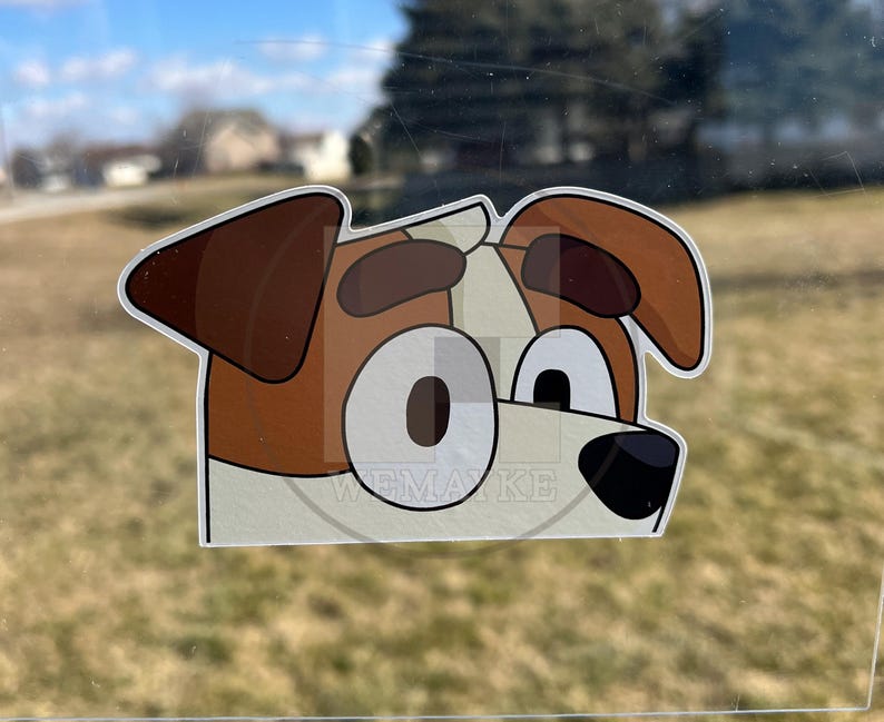 May include: A die-cut vinyl sticker featuring a cartoon dog head with brown ears, brown eyebrows, and large white eyes with dark pupils. The dog has a white muzzle and a black nose. The text WEMAYKE is visible in a faint watermark across the center.