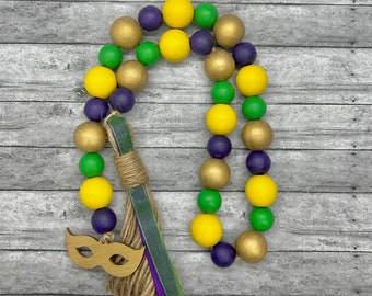 May include: A Mardi Gras garland made of wooden beads in yellow, green, and purple colors. The garland has a wooden mask charm and a burlap tassel.