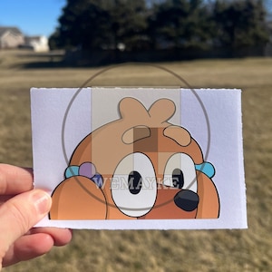 May include: A rectangular white card featuring a cartoon illustration of a brown dog character with large white eyes, a black nose, and small blue and purple circular accents on the sides, with the text WEMAYKE printed in white across the center.
