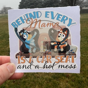 May include: A white sticker with a blue, orange, and brown cartoon illustration of two dogs in car seats. The text reads "Behind every Mama is a car seat and a hot mess."