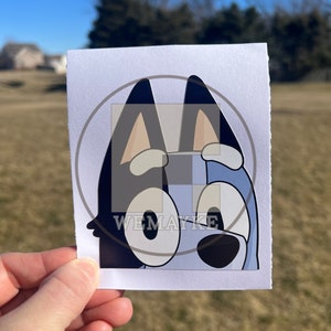 May include: A rectangular white sticker featuring a cartoon illustration of a blue dog character with large eyes, pointed ears, and a black nose. The sticker has a semi-transparent circular overlay with the text WEMAYKE printed in white letters.