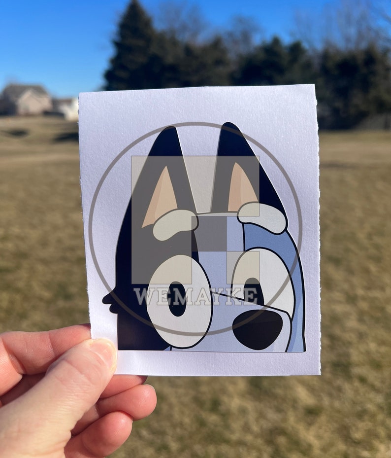 May include: A rectangular white sticker featuring a cartoon illustration of a blue dog character with large eyes, pointed ears, and a black nose. The sticker has a semi-transparent circular overlay with the text WEMAYKE printed in white letters.