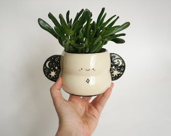 May include: A cream-colored ceramic planter with a smiling face and a star design. The planter has two black handles with a moon and star pattern. It holds a green succulent plant.