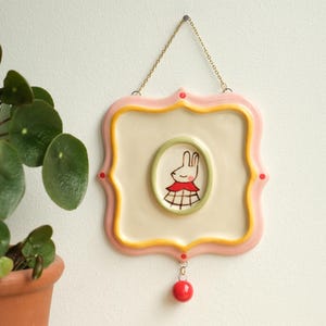 May include: A square ceramic wall plaque with scalloped edges, featuring a cream-coloured centre, a pink and yellow border, and a central oval frame containing a hand-painted illustration of a rabbit wearing a red collar and plaid dress.