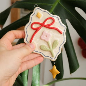 Pode incluir: Um ornamento de cerâmica cor de creme com uma borda ondulada delineada em azul. Apresenta um laço vermelho em relevo, um pequeno diamante amarelo e uma flor rosa com folhas verdes. Um pequeno pendente amarelo em forma de estrela pende de um anel.