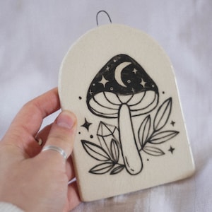 May include: A white ceramic wall hanging with a black and white illustration of a mushroom with a crescent moon and stars on the cap. The mushroom is surrounded by leaves and a crystal.