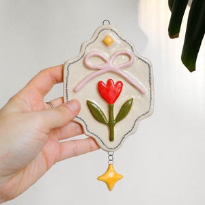 May include: A cream-coloured ceramic wall hanging with a scalloped edge, featuring a raised red tulip with green leaves, a light pink bow, and a small yellow diamond accent. A yellow four-pointed star charm hangs from the bottom by a metal chain.