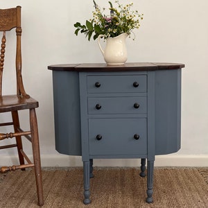 May include: A blue painted wooden table with three drawers and turned legs. The table has a dark wood top and is decorated with a vase of flowers.