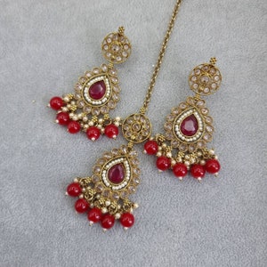 Peut inclure: Un ensemble de bijoux de couleur or antique comprenant un serre-t&ecirc;te et des boucles d'oreilles assorties. Chaque pi&egrave;ce comporte une pierre pr&eacute;cieuse rouge fonc&eacute; en forme de goutte d'eau, entour&eacute;e de petites perles blanches et de cristaux transparents. Les boucles d'oreilles et le serre-t&ecirc;te sont orn&eacute;s de grappes de petites perles rouges.