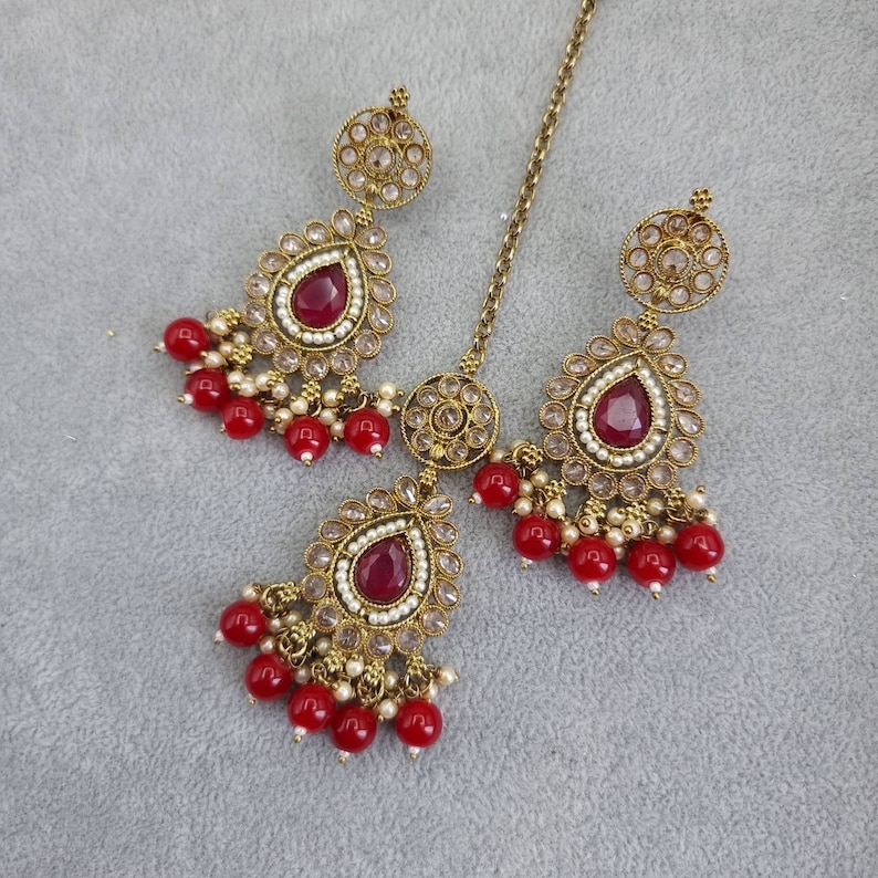 Peut inclure: Un ensemble de bijoux de couleur or antique comprenant un serre-t&ecirc;te et des boucles d'oreilles assorties. Chaque pi&egrave;ce comporte une pierre pr&eacute;cieuse rouge fonc&eacute; en forme de goutte d'eau, entour&eacute;e de petites perles blanches et de cristaux transparents. Les boucles d'oreilles et le serre-t&ecirc;te sont orn&eacute;s de grappes de petites perles rouges.