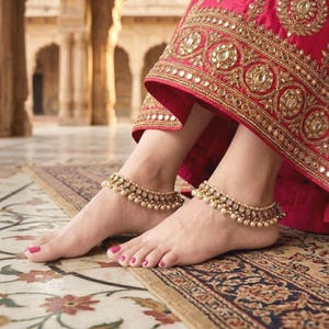 May include: Gold-toned anklets with pearl detailing are worn on the ankles. The anklets are paired with a red and gold embroidered garment. The image is taken indoors on a patterned rug.