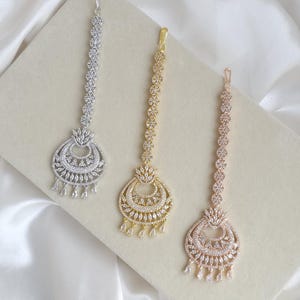 Op de afbeelding: Drie maang tikka hoofd sieraden in zilver-, goud- en roségoudtinten. Elk stuk heeft een lange ketting van cirkelvormige bloemmotieven die leidt naar een halvemaanvormige hanger versierd met heldere kristallen en kleine druppelvormige steentjes.