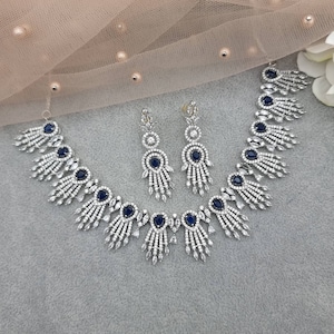 May include: A silver-toned necklace and earring set. The necklace and earrings feature a design with clear and dark blue stones. The necklace has a series of dark blue oval stones and fan-shaped accents. The earrings have a similar design.