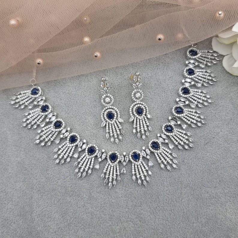 May include: A silver-toned necklace and earring set. The necklace and earrings feature a design with clear and dark blue stones. The necklace has a series of dark blue oval stones and fan-shaped accents. The earrings have a similar design.