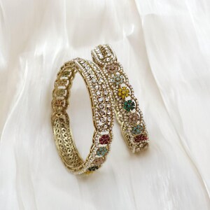 May include: Two gold-toned bangles with intricate designs and colorful gemstones. One bangle features a floral pattern with red, yellow, green, and blue accents. The other bangle has a detailed, openwork design. The bangles rest on a white, textured surface.