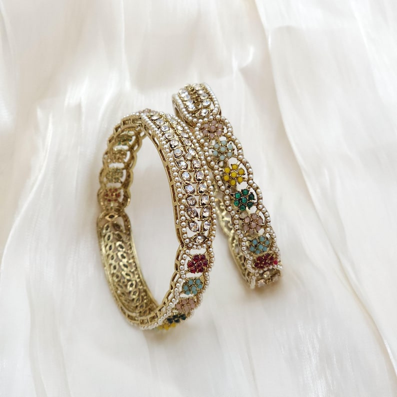 May include: Two gold-toned bangles with intricate designs and colorful gemstones. One bangle features a floral pattern with red, yellow, green, and blue accents. The other bangle has a detailed, openwork design. The bangles rest on a white, textured surface.
