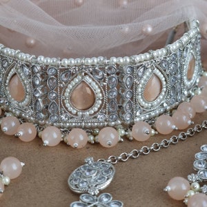 Peut inclure: Un ensemble de bijoux de mari&eacute;e argent&eacute; comprenant un collier ras du cou, des boucles d'oreilles et un bijou de t&ecirc;te. Le ras du cou est orn&eacute; de pierres en forme de goutte de couleur p&ecirc;che, de perles blanches et de cristaux transparents.