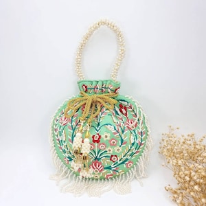 May include: A small, round, green drawstring bag with a floral embroidery design. The bag has a white pearl handle and a white pearl fringe around the bottom. The bag is decorated with gold beads and tassels.