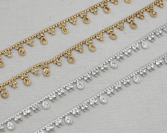 May include: Close-up of gold and silver decorative trim. The gold trim features small, round accents and teardrop-shaped embellishments. The silver trim has teardrop-shaped clear crystal accents. The trim is arranged diagonally.