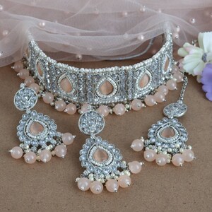 Peut inclure: Un ensemble de bijoux de mari&eacute;e argent&eacute;s comprenant un collier ras du cou, des boucles d'oreilles et un serre-t&ecirc;te. L'ensemble comprend des pierres en forme de goutte d'eau de couleur p&ecirc;che, de petites perles blanches et des perles de couleur p&ecirc;che. Les bijoux sont pr&eacute;sent&eacute;s sur un fond neutre.