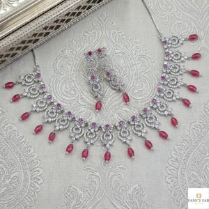 May include: A silver-toned necklace and matching earrings set. The jewelry features a floral design with clear stones and dangling oval-shaped, ruby-colored beads. The set is displayed on a patterned, light-colored surface.