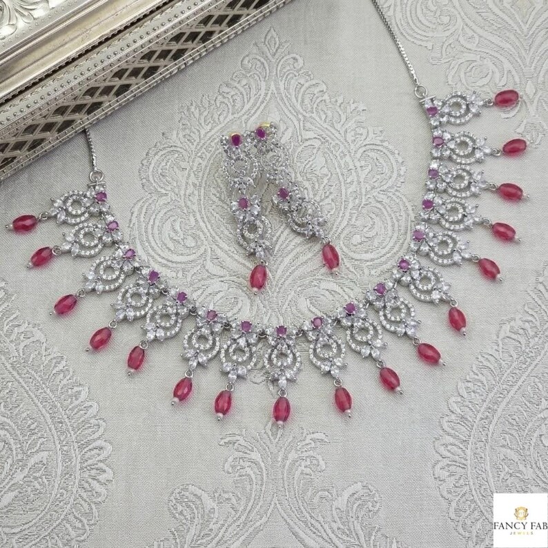May include: A silver-toned necklace and matching earrings set. The jewelry features a floral design with clear stones and dangling oval-shaped, ruby-colored beads. The set is displayed on a patterned, light-colored surface.