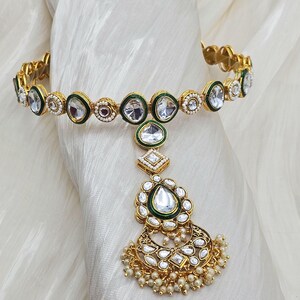 May include: Gold-toned headpiece with clear, faceted stones and green enamel accents. The design includes a circular band, a central pendant with a teardrop-shaped stone, and a dangling, ornate floral element with small pearl beads.