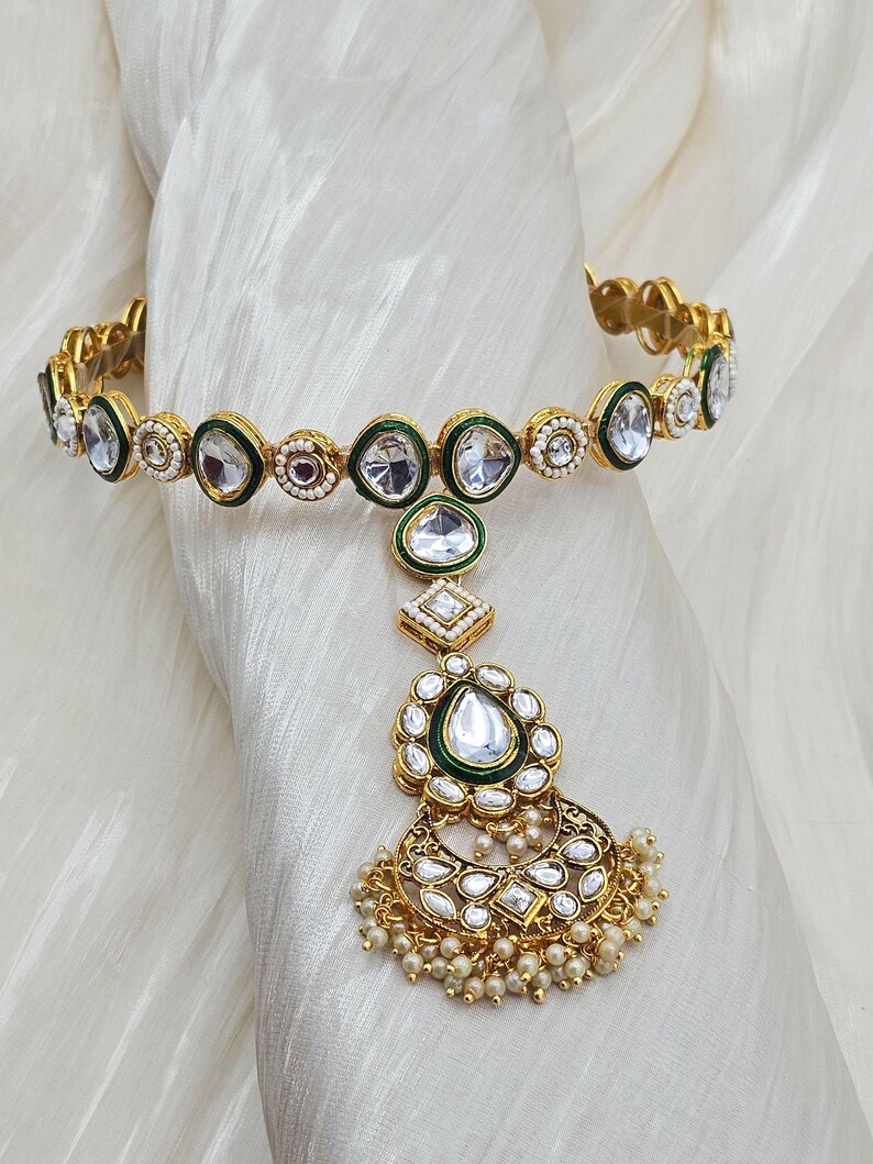 May include: Gold-toned headpiece with clear, faceted stones and green enamel accents. The design includes a circular band, a central pendant with a teardrop-shaped stone, and a dangling, ornate floral element with small pearl beads.
