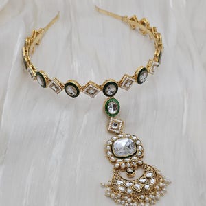 May include: Gold-toned headband with geometric and circular accents, featuring clear and green gemstones. A dangling pendant with a large, clear gemstone and pearl accents completes the design. The jewelry is set against a white, textured background.
