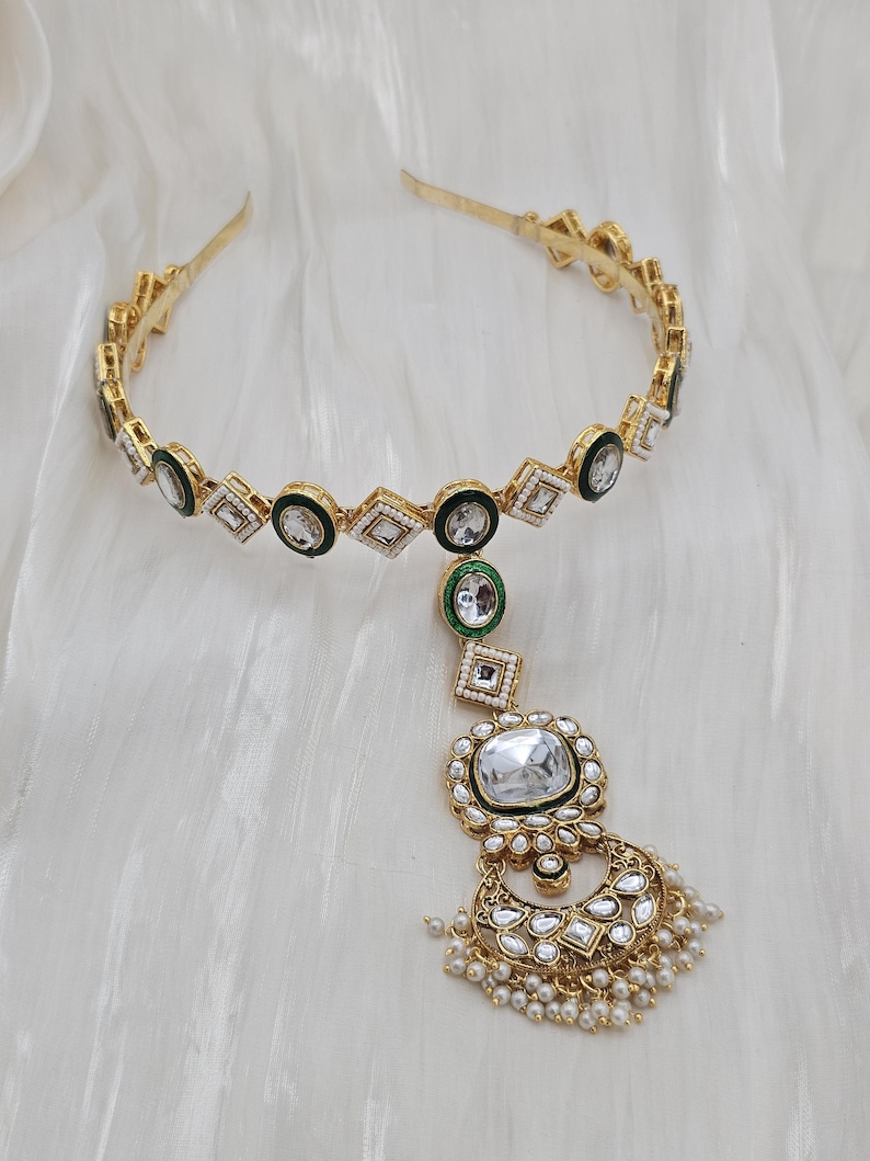 May include: Gold-toned headband with geometric and circular accents, featuring clear and green gemstones. A dangling pendant with a large, clear gemstone and pearl accents completes the design. The jewelry is set against a white, textured background.