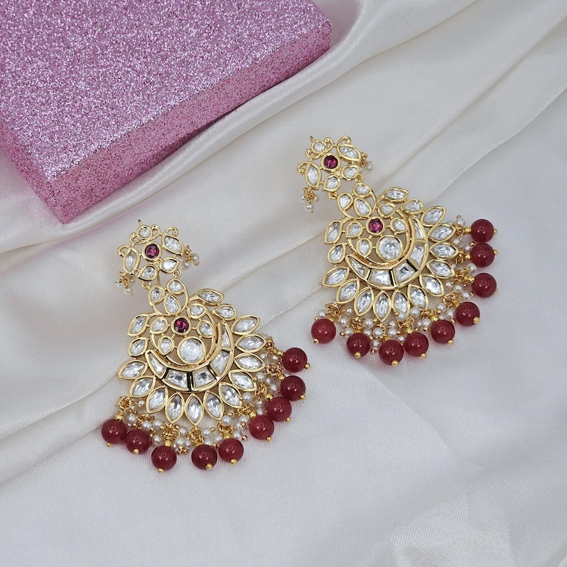 May include: A pair of gold-toned chandelier earrings with clear and red gemstones. The earrings have a floral design with dangling red beads and small white pearls. The earrings are displayed on a white fabric surface.