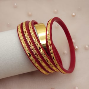May include: A collection of red bangles with gold decorative detailing. The bangles are arranged together, with a gold bangle in the middle. The background is a soft, pale pink colour.