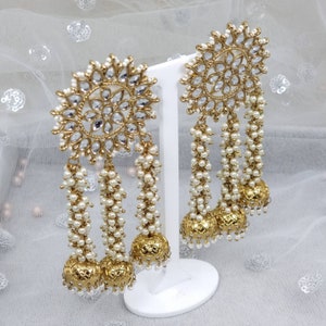 May include: A pair of gold tone earrings with a large, round, clear crystal design and three strands of white pearl beads hanging from each earring.