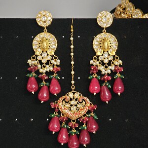 Pu&ograve; includere: Un set di gioielli da sposa indiani in oro e rosso con un ciondolo a forma di lacrima e orecchini abbinati. Il set presenta disegni intricati, perle bianche e pietre preziose rosse e verdi.