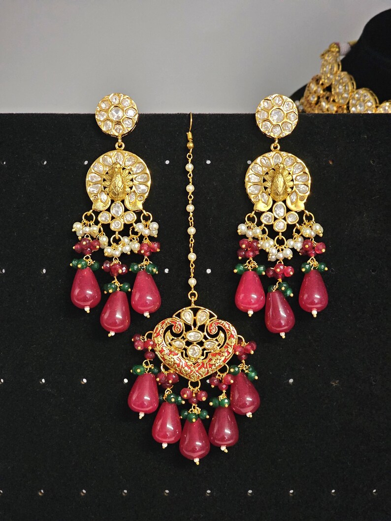 Pu&ograve; includere: Un set di gioielli da sposa indiani in oro e rosso con un ciondolo a forma di lacrima e orecchini abbinati. Il set presenta disegni intricati, perle bianche e pietre preziose rosse e verdi.