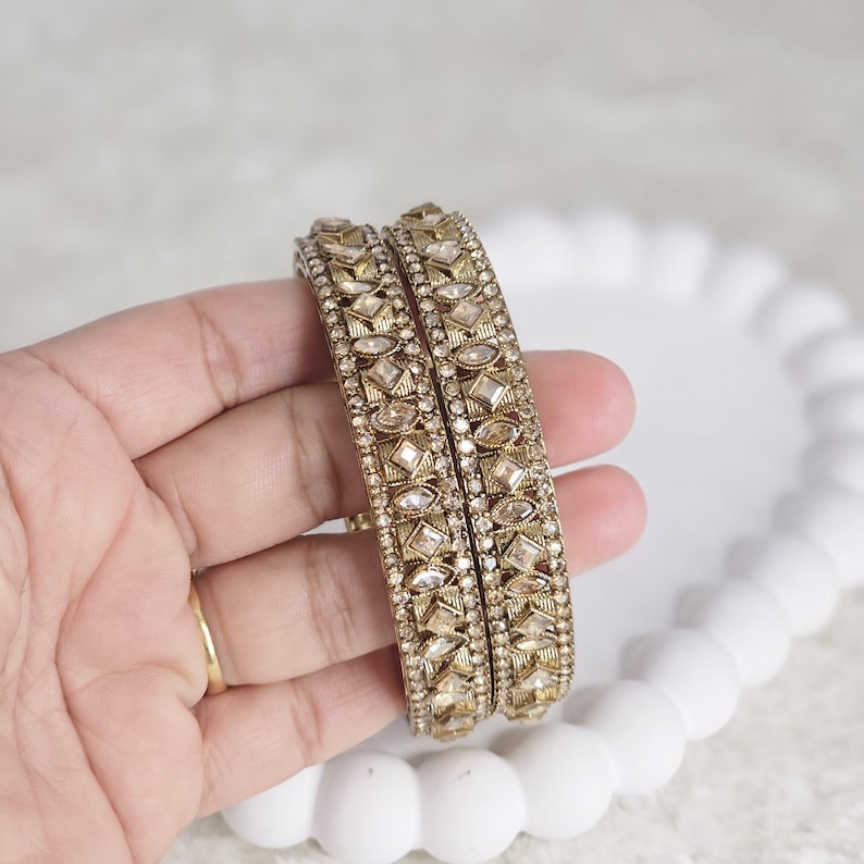 May include: Two gold-toned bangles with intricate geometric patterns and embedded clear gemstones. The bangles are held in a hand, showcasing their detailed design and sparkling accents. The jewelry is set against a soft, neutral background.