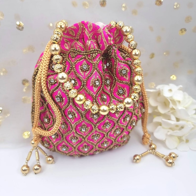 Puede incluir: Una bolsa de cord&oacute;n rosa con bordados y adornos dorados. La bolsa tiene un cord&oacute;n de cord&oacute;n de cuentas doradas y acentos de borla.