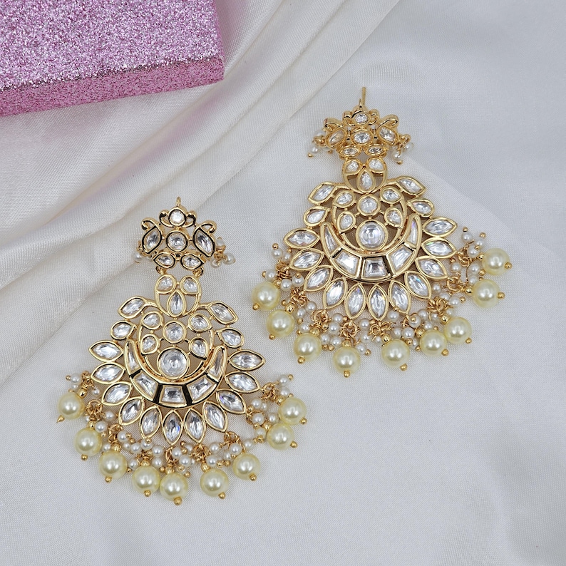 May include: A pair of gold-toned earrings with a floral design, featuring clear, faceted stones and dangling faux pearls. The earrings have a chandelier style and are set against a white fabric background.