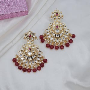 May include: Gold-toned chandelier earrings with clear and ruby-colored stones and small white pearls. The earrings have a floral design and are adorned with ruby-colored beads. The earrings are displayed on a white, silky fabric.