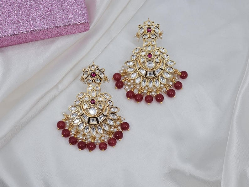 May include: Gold-toned chandelier earrings with clear and ruby-colored stones and small white pearls. The earrings have a floral design and are adorned with ruby-colored beads. The earrings are displayed on a white, silky fabric.