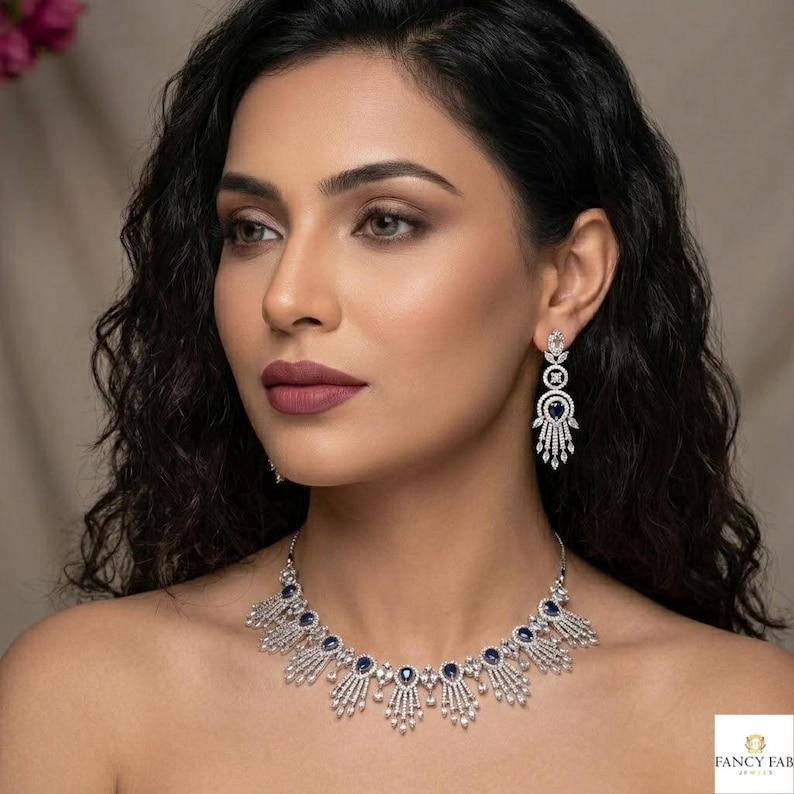 Elegant silver-toned bridal jewelry set featuring a necklace and matching drop earrings. The design showcases teardrop-shaped sapphire blue stones surrounded by intricate cubic zirconia or diamond-style fringes in a sunburst pattern. Modeled on a woman with wavy dark hair and soft glam makeup, highlighting how the pieces sit on the neckline and ears. Ideal for weddings, formal galas, or as a luxury gift for her.