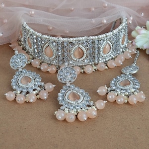 Peut inclure: Un ensemble de bijoux de mari&eacute;e argent&eacute;s comprenant un collier ras du cou, des boucles d'oreilles et un bijou de t&ecirc;te. L'ensemble comprend des pierres en forme de goutte d'eau de couleur p&ecirc;che, de petites perles et des pampilles de perles p&ecirc;che. Les bijoux sont pr&eacute;sent&eacute;s sur un fond neutre.